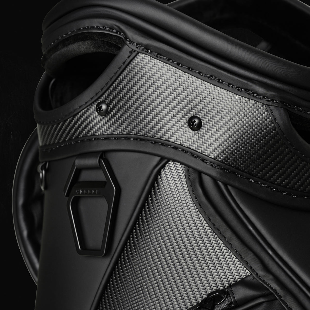 Close-up of the top of a black VESSEL golf bag with carbon fiber panels, matte black trim and a black metal ring engraved with VESSEL
