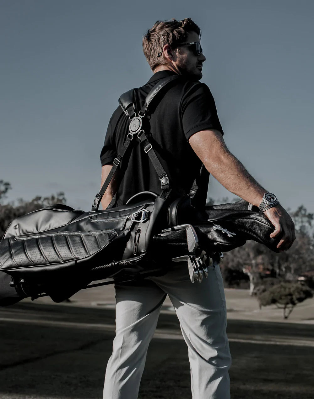Golfer in a black polo shirt and sunglasses on a golf course, carrying a black VESSEL stand bag on a double shoulder strap