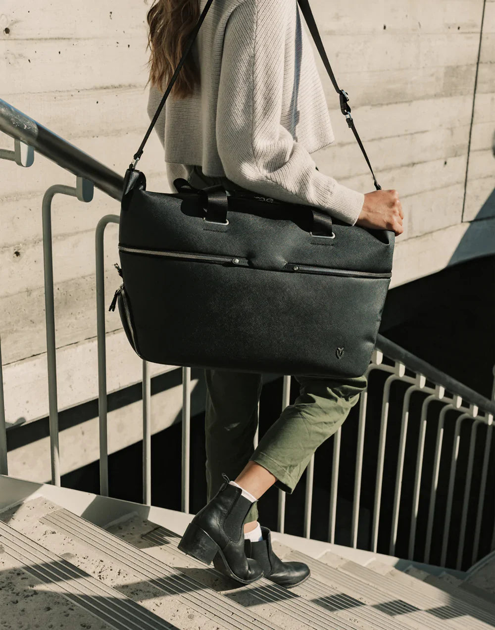 Woman in a cream knit sweater walking down an outdoor concrete staircase with a black VESSEL duffel bag on her shoulder