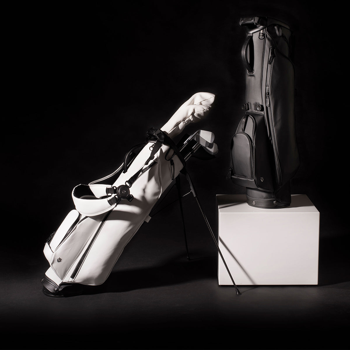 A white VESSEL stand bag with clubs propped on its legs beside a black VESSEL bag standing on a white plinth, in a dark studio