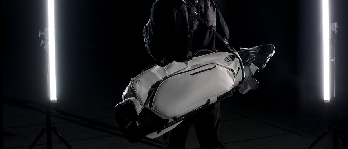 Golfer dressed in black carrying a white VESSEL stand bag with clubs on a double shoulder strap, in a dark studio between two tall light tubes