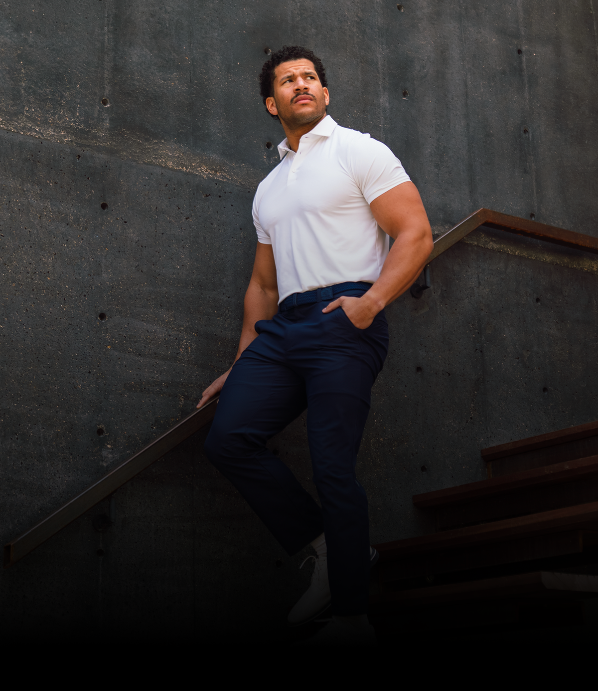 Man in a white polo shirt and navy trousers leaning on the handrail of an outdoor concrete staircase