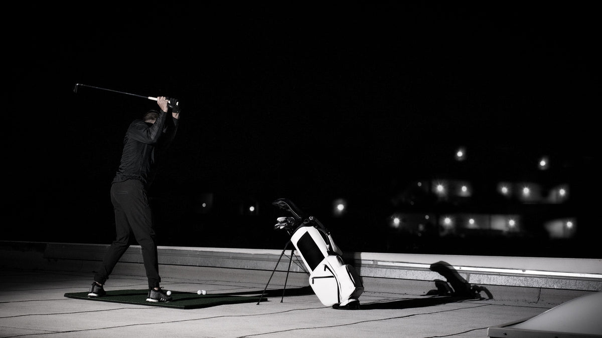 Golfer at the top of his backswing on a rooftop practice mat at night, with a white golf stand bag beside him and city lights behind